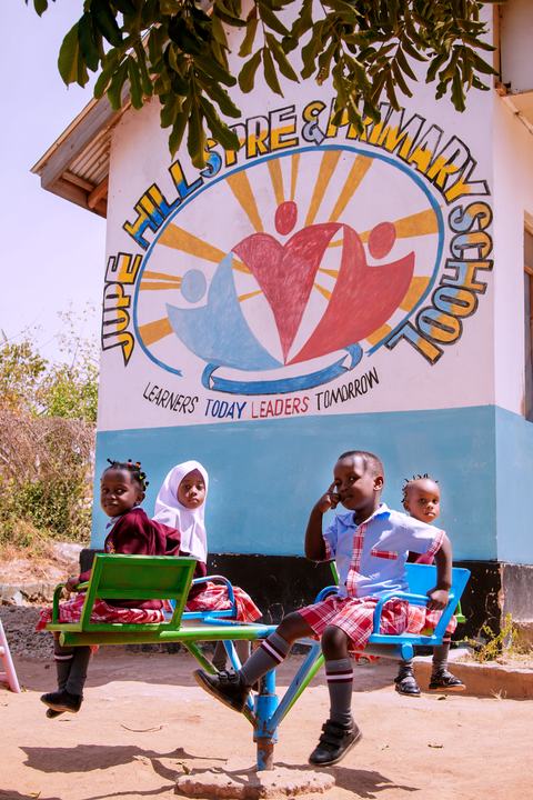 Young children sitting on playground seesaws in front of the Jupe Hills school mural